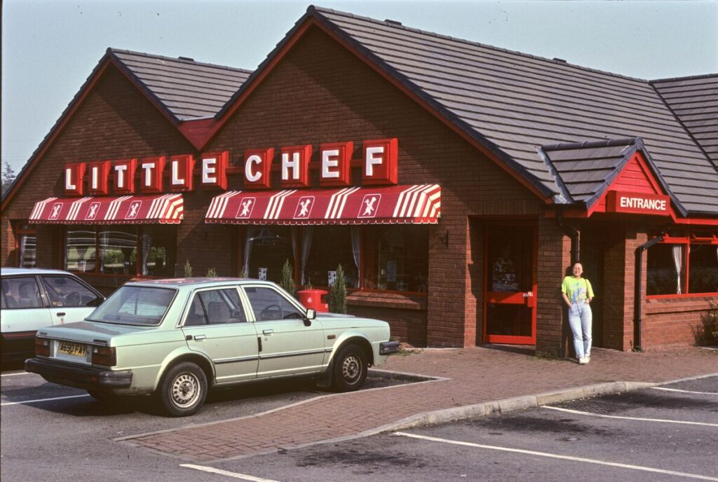 photograph of a 'Little Chef' restaurant with cars parked outside and a woman standing near the entrance